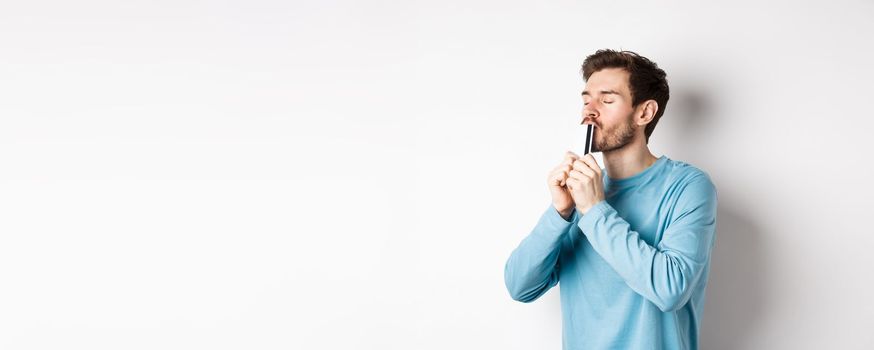 Young man kissing plastic credit card with satisfied face, standing over white background in blue casual shirt.