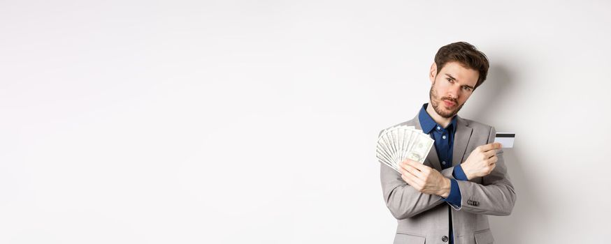 Cool young rich man in suit showing dollar bills and plastic credit card, making money, standing on white background.