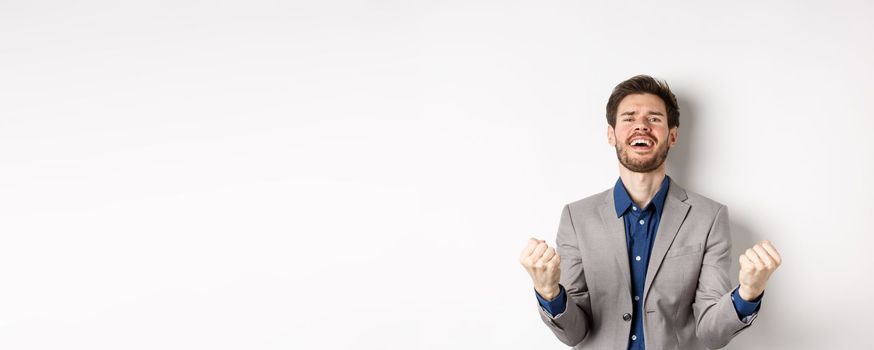 Excited man winning in cazino and celebrating, making fist pump and shouting yes with happy expression, achieve goal, triumphing, standing on white background.