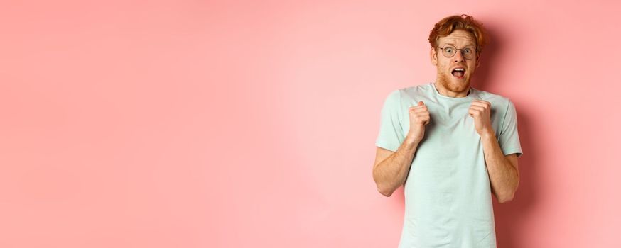 Portrait of scared redhead guy in glasses staring startled at camera, press hands to body and scream of fear, standing over pink background.