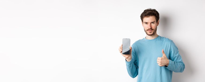 Young man showing empty smartphone screen and thumb up, recommend mobile application or online shopping offer, standing over white background.