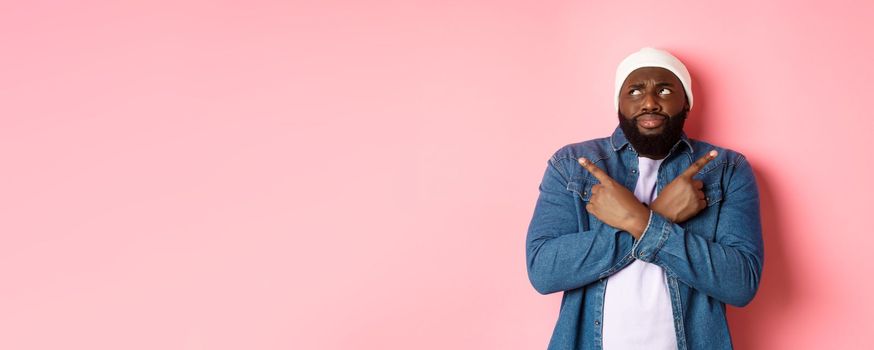 Confused Black man with beard, making choice, pointing fingers sideways and looking puzzled, standing over pink background.