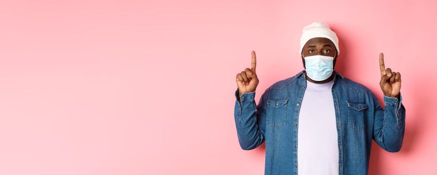 Coronavirus, lifestyle and global pandemic concept. Doubtful african-american man in face mask pointing fingers up, showing copy space, pink background.