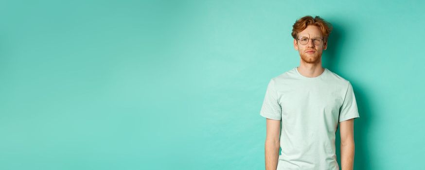 Confused young redhead man in glasses staring at camera puzzled, squinting perplexed, cant understand something, standing over mint background.