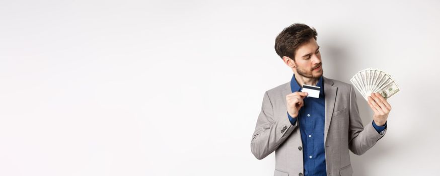 Successful man showing plastic credit card and looking at dollar bills fan, making money deposit, standing on white background.