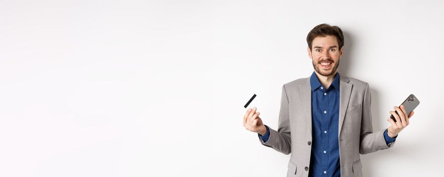 Online shopping. Excited man making money, smiling amazed, holding smartphone and credit card, standing against white background in suit.