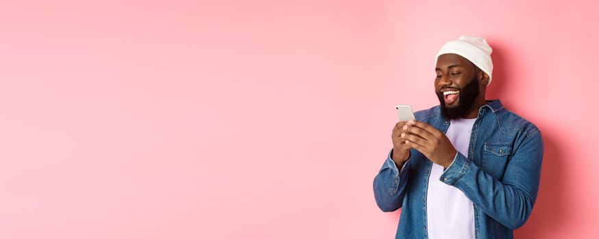 Technology and online shopping concept. Happy Black bearded man reading message and smiling, using smartphone against pink background.