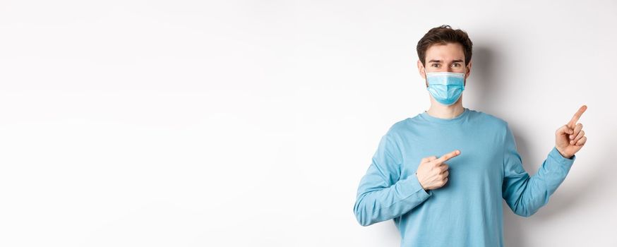 Coronavirus, health and quarantine concept. Handsome caucasian man in face mask pointing fingers right, showing advertisement, standing over white background.