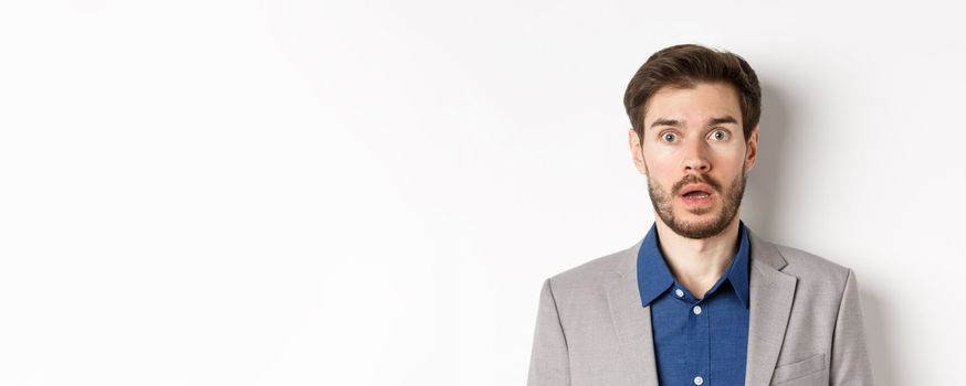 Close up portrait of shocked caucasian man in suit drop jaw, looking speechless at camera, white background.