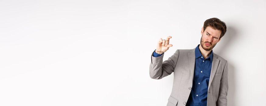 Businessman trying to see something tiny, showing small thing size with fingers, standing in suit on white background.