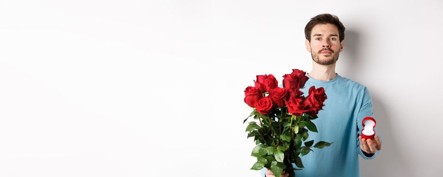 Valentines and relationship. Romantic man boyfriend holding red roses and showing engagement ring, making a proposal on lovers day, standing over white background.