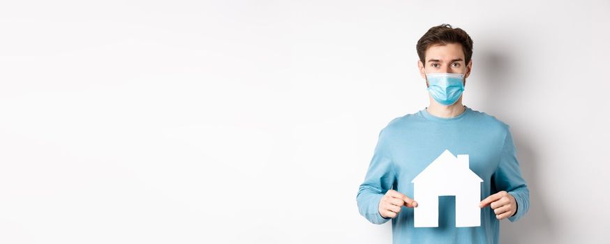 Covid and real estate concept. Man searching for flat, showing paper house cutout, wearing medical mask, standing on white background.