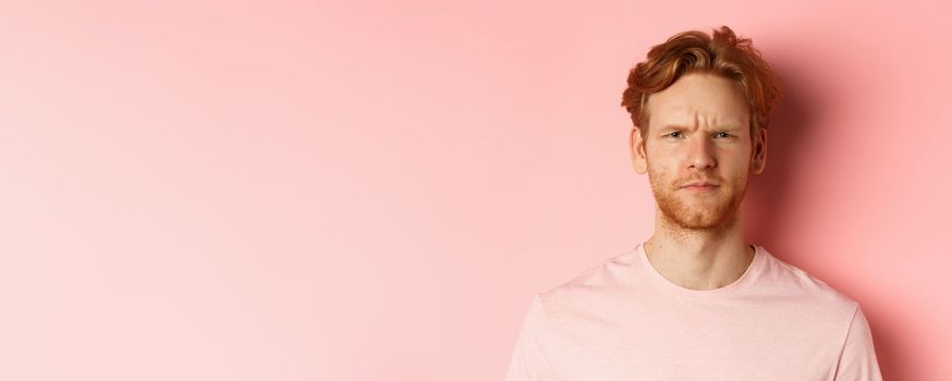 Close up of suspicious redhead guy frowning, staring at camera doubtful, express disapproval, standing over pink background.