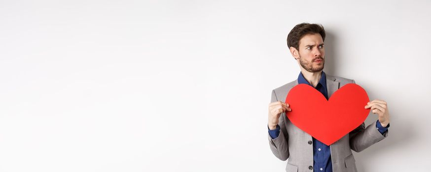 Confused man in suit holding big red heart and looking right, searching for love on Valentines day, standing over white background.