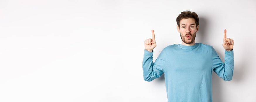 Surprised young man showing top advertisement, pointing up and saying wow at camera, standing over white background.