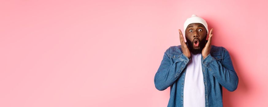 Shocked and impressed Black man staring at camera with complete disbelief, saying wow, standing in beanie and hipster shirt over pink background.