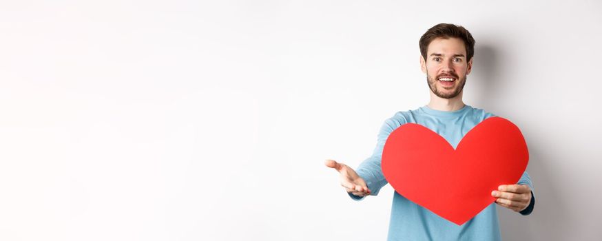 Handsome man in love making confession to you, pointing hand at camera, holding big red heart cutout on valentines day, singing romantic serenade, standing over white background.