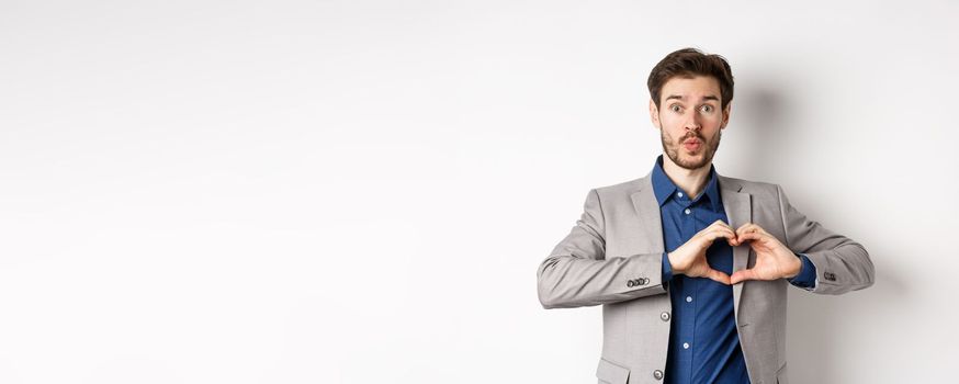 Handsome man looking silly, showing heart love you sign and pucker lips, waiting for kiss from lover, standing in suit on white background.