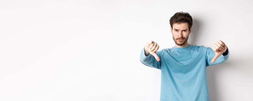 Skeptical handsome guy with beard, showing thumbs down in disapproval, dislike something bad, standing over white background.