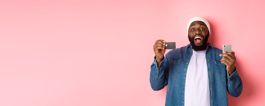 Online shopping. Happy bearded african-american man smiling, showing credit card and making purchase on smartphone, standing over pink background.