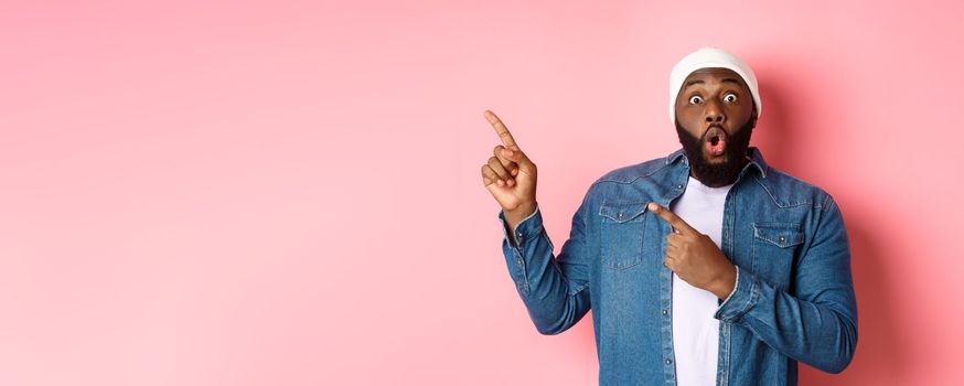 Amazed Black guy showing awesome promo offer, staring at camera excited and smiling, pointing fingers at upper left corner banner, pink background.