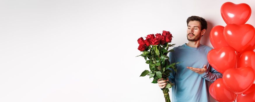 Romantic guy showing bouquet of red roses, pointing at flowers and standing near hearts balloons on Valentines day, prepare gifts for his lover, white background.