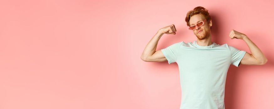 Confident young man with red hair, wearing summer sunglasses and t-shirt, showing strong and fit body muscles, flex biceps and staring cool at camera, pink background. Workout and gym concept