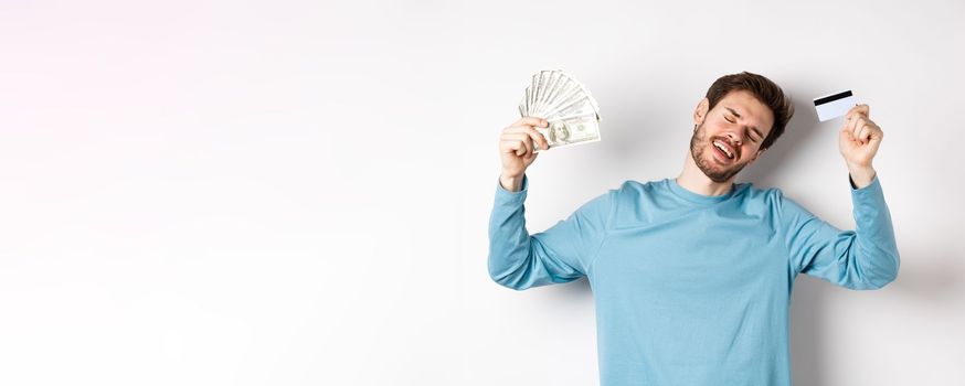 Handsome man dancing with money and plastic credit card, standing in casual clothes over white background.