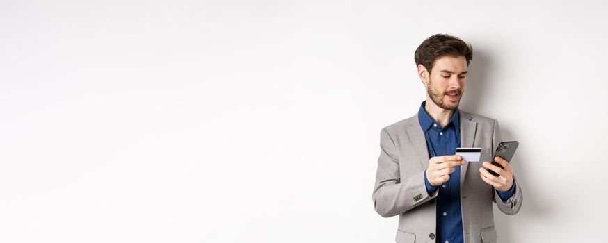 Online shopping. Smiling businessman paying with credit card on smartphone, sending money, standing in suit on white background.