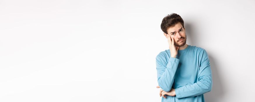 Sad young man looking frustrated, touching temple of head and frowning uneasy, standing against white background.