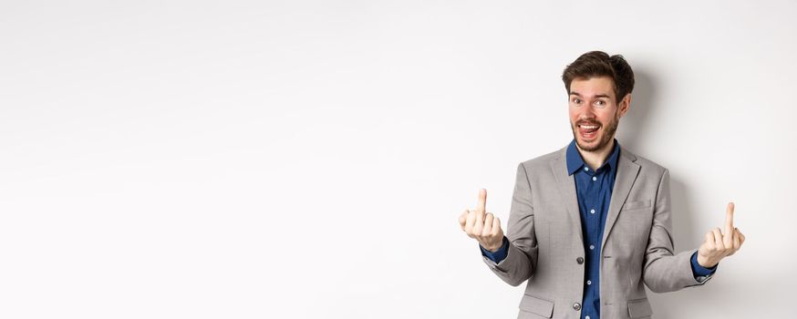 Rude ignorant guy in business suit showing middle fingers and tongue, smiling while mocking people, fuck you gesture, standing on white background.