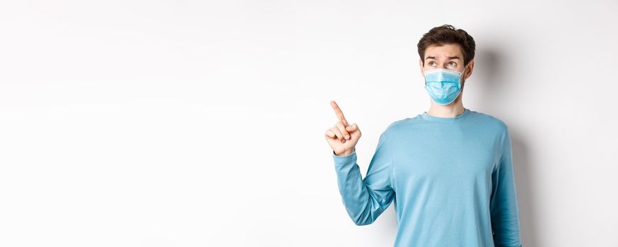 Coronavirus, health and quarantine concept. Curious guy in medical mask, pointing and looking at upper left corner banner, standing on white studio background.