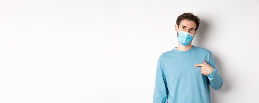 Covid-19, health and quarantine concept. Smiling caucasian man in medical mask pointing at himself, volunteering, standing over white background.