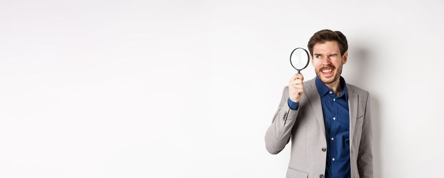 Funny guy in suit investigating or searching for something, looking through magnifying glass, standing on white background.