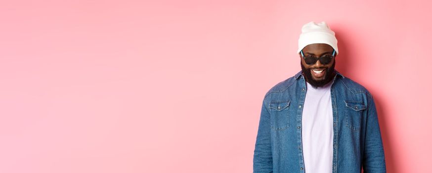 Cool african-american hipster man in beanie and sunglasses, smiling pleased and looking at camera, standing confident over pink background.