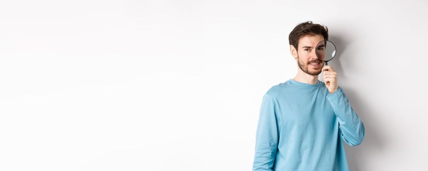 Smiling young man detective looking through magnifying glass, searching for promo offer, standing on white background.