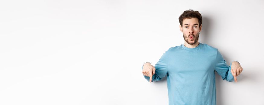 Amazed young man with beard, standing in casual blue shirt, saying wow and pointing down, look here gesture, standing over white background.