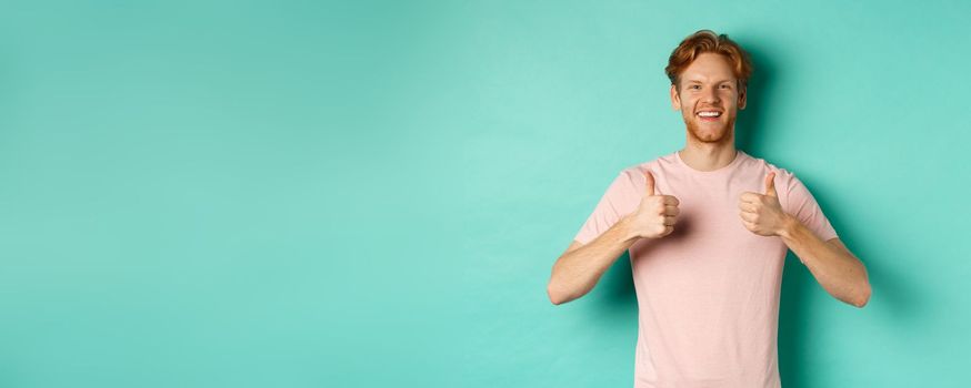 Cheerful bearded man with red hair showing thumbs-up, like and approve something, praising promo, standing against turquoise background.