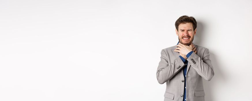 Crying man in suit holding hands on neck and look distressed, feel sick with sore throat, standing on white background.