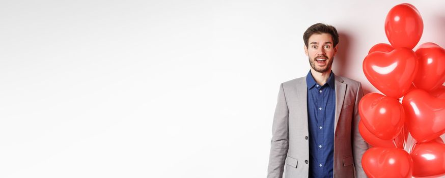 Lovers day. Excited handsome man in suit standing near red hearts balloons, raising eyebrows and looking surprised, standing over white background.