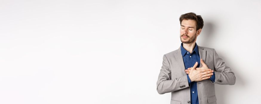 Romantic bearded man in stylish suit holding hands on heart, close eyes and remember something with tender feelings, standing nostalgic on white background.