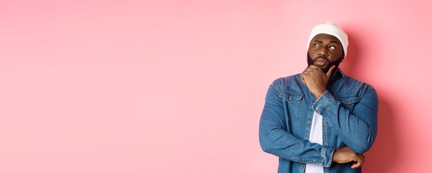 Serious thoughtful african american male model making decision, thinking and staring at upper left corner, choosing or pondering, standing over pink background.