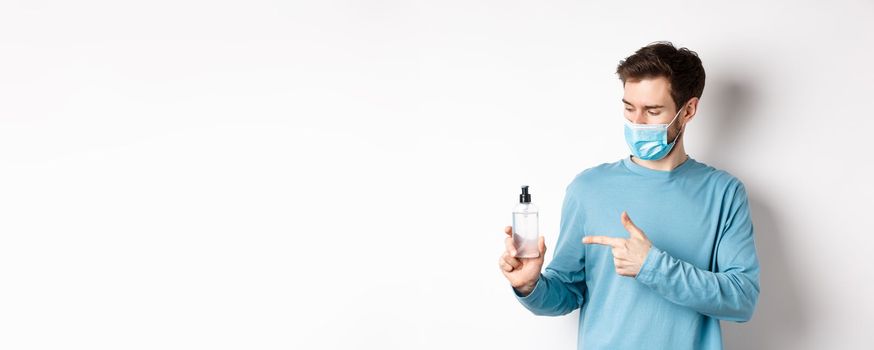 Covid-19, health and quarantine concept. Young man pointing and looking at hand sanitizer bottle, showing good antiseptic, white background.