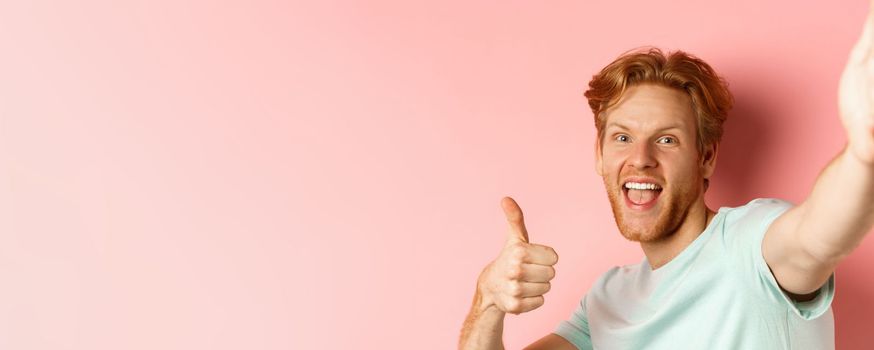 Excited redhead man tourist taking selfie and showing thumbs-up, holding camera with stretched out hand, standing over pink background.