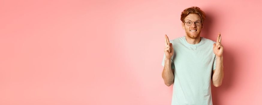 Worried redhead man waiting for results, expecting something with fingers crossed, biting finger and looking at something risky, standing over pink background.