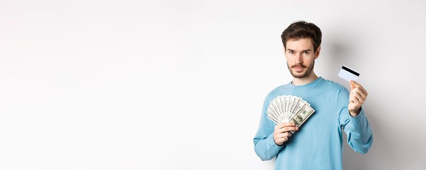 Caucasian man holding money and showing plastic credit card, standing in blue sweatshirt on white background.