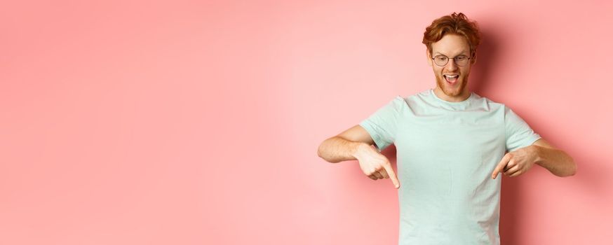 Handsome caucasian man with red messy hair and glasses, pointing fingers and looking down at promo offer, smiling pleased, standing over pink background.