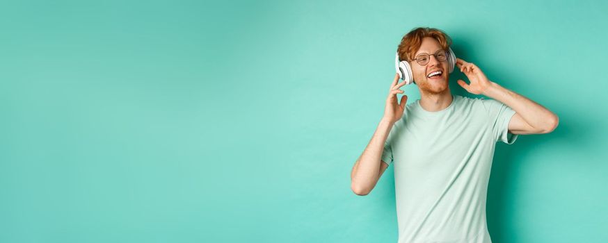 Lifestyle concept. Happy young man with ginger hair dancing and having fun, listening music on wireless headphones and smiling pleased, turquoise background.