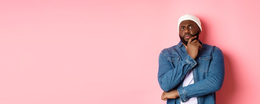 Doubtful african american man staring at camera with disbelief, standing skeptical and thinking, pink background.