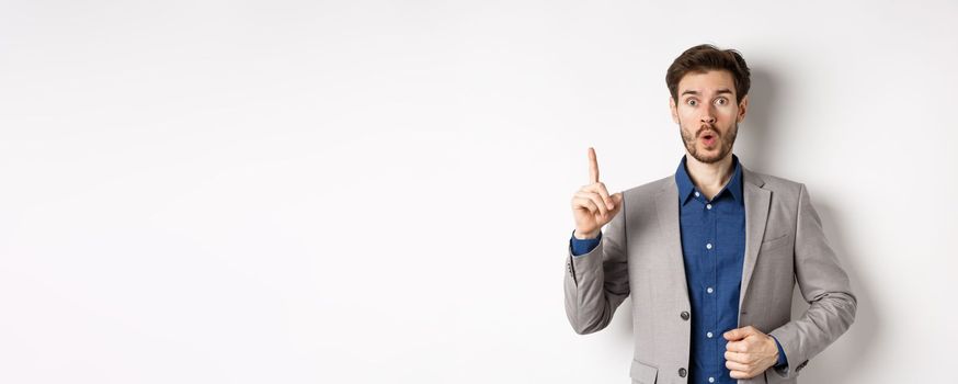 Excited businessman in suit pitching good idea, raising finger in eureka sign and looking amused, saying suggestion, standing on white background.